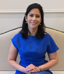 Woman sitting on a beige couch wearing a blue dress, smiling.